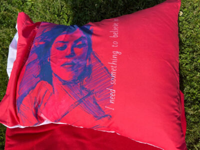 A pillow showing a blue sketch of a woman on a red background. Text reads "I need something to believe in."