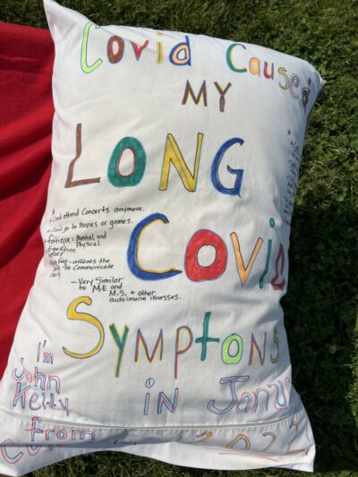 Pillow reads "COVID caused by Long COVID symptoms in January, 2022. I'm John Kelty." Other text is obscured, and some is written in smaller print elsewhere on the pillowcase.