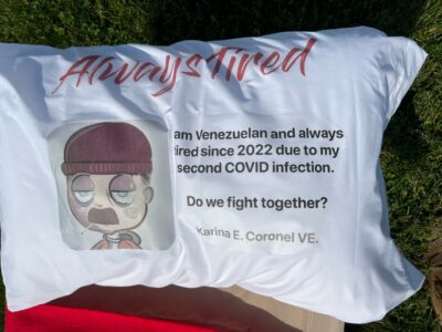 A pillow showing a cartoon of a mustached man with a beanie cap pulled just above his hooded eyes. A cigarette hangs loosely from his mouth. Text reads "Always Tired. I am Venezuelan and always tired since 2022 due to my second COVID infection. Do we fight together? Karina E. Coronel VE."